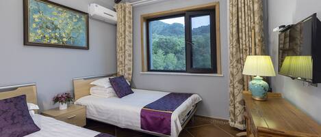 A hotel room with two beds, a window with a view of greenery, a mounted TV, and a bedside table with a lamp.