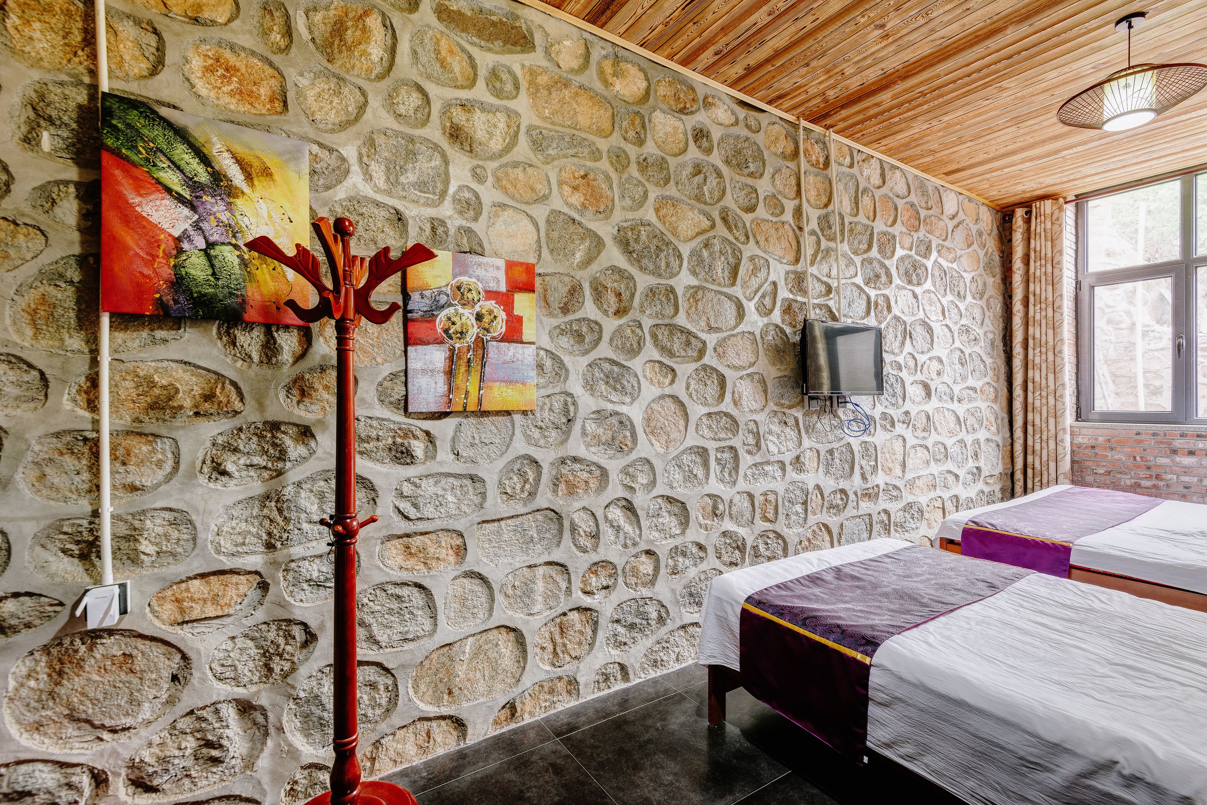 Une pièce aux murs de pierre, au plafond en bois, avec deux lits dotés de draps violets et blancs, un téléviseur accroché au mur et un tableau sur le côté gauche.