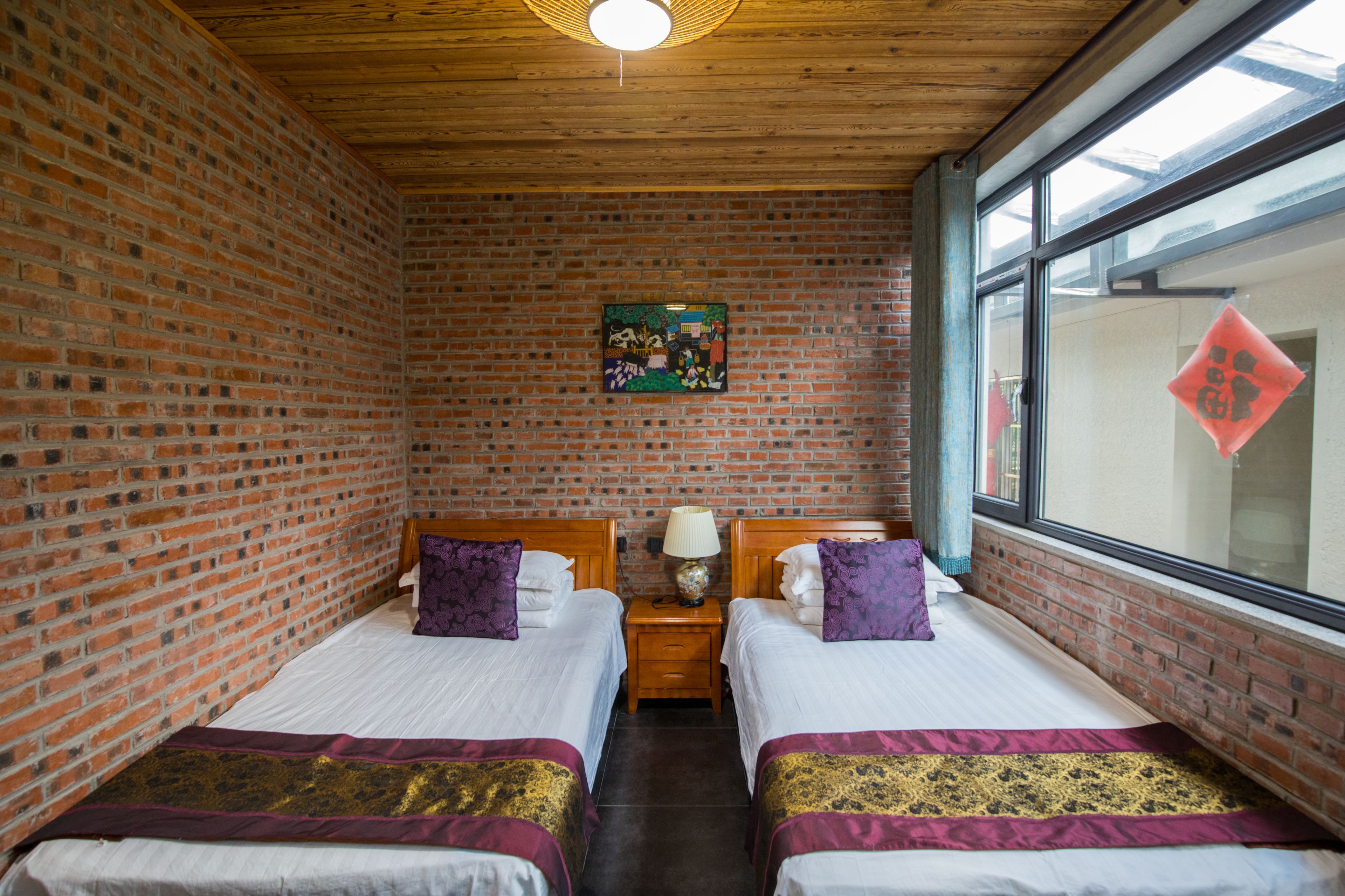 A room with two beds, a wooden ceiling, brick walls, a window with a view, and a painting on the wall.