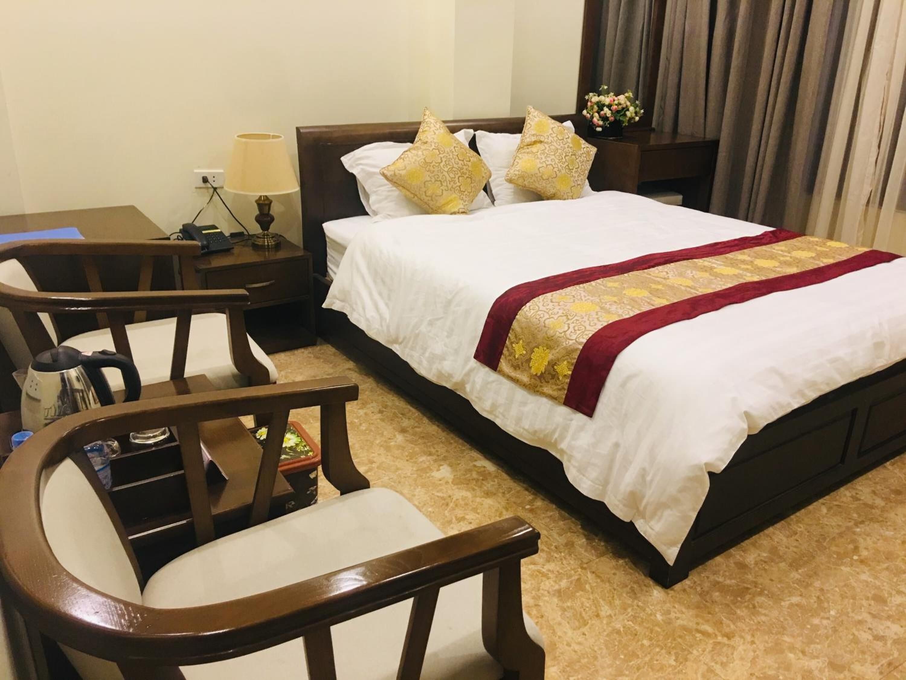 A hotel room with a bed, two chairs, a nightstand with a lamp, and a vase of flowers.