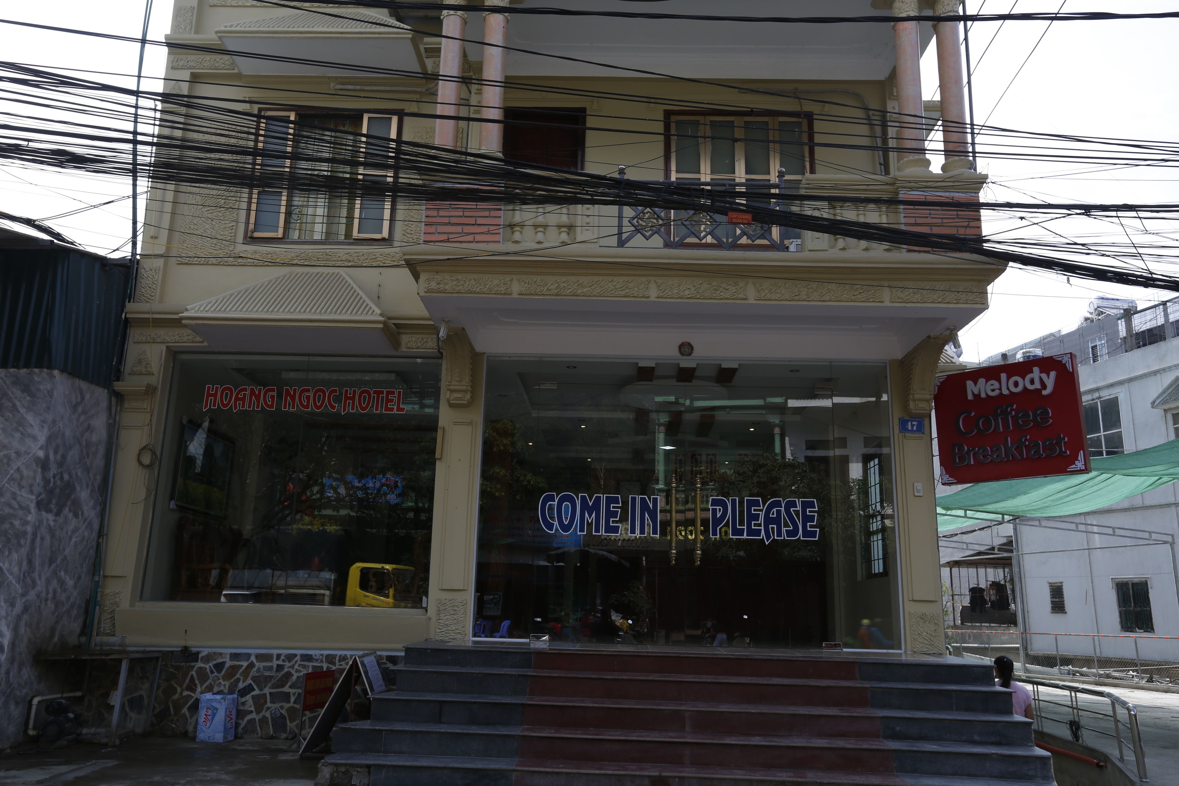 Una entrada de hotel con un cartel que dice 'COME IN PLEASE', un cartel de 'Melody Coffee Breakfast' y un cartel de 'HOANG NGOC HOTEL'.