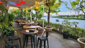 An outdoor seating area with tables and chairs, potted plants, and a view of a river with boats.