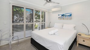 A bedroom with a bed, a ceiling fan, a nightstand, a chair, and a window with blinds. - Illawong Apartments (Mooloolaba)