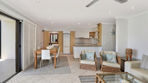 A modern apartment with a dining area, two wicker sofas, a glass coffee table, and a kitchen in the background. - Illawong Apartments (Mooloolaba)