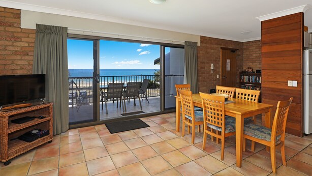 A room with a dining table and chairs, a television on a stand, and a view of the beach through a sliding glass door. - Illawong Apartments (Mooloolaba)