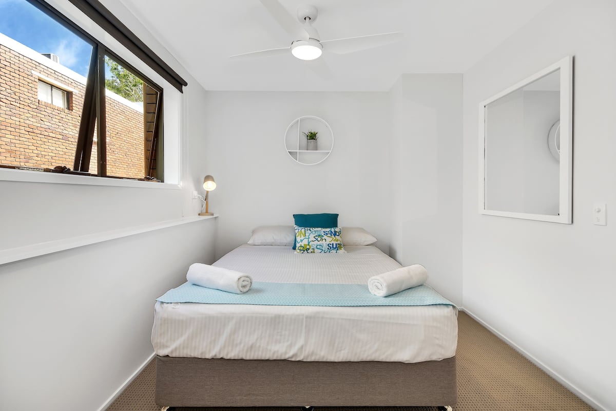 A neatly made bed with white bedding and a blue throw, a round wall shelf with a plant, and a ceiling fan.