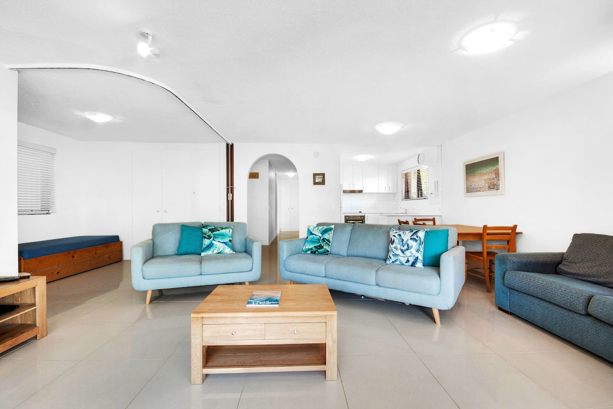 A modern living room with two light blue sofas, a wooden coffee table, and a dining area with a wooden table and chairs.