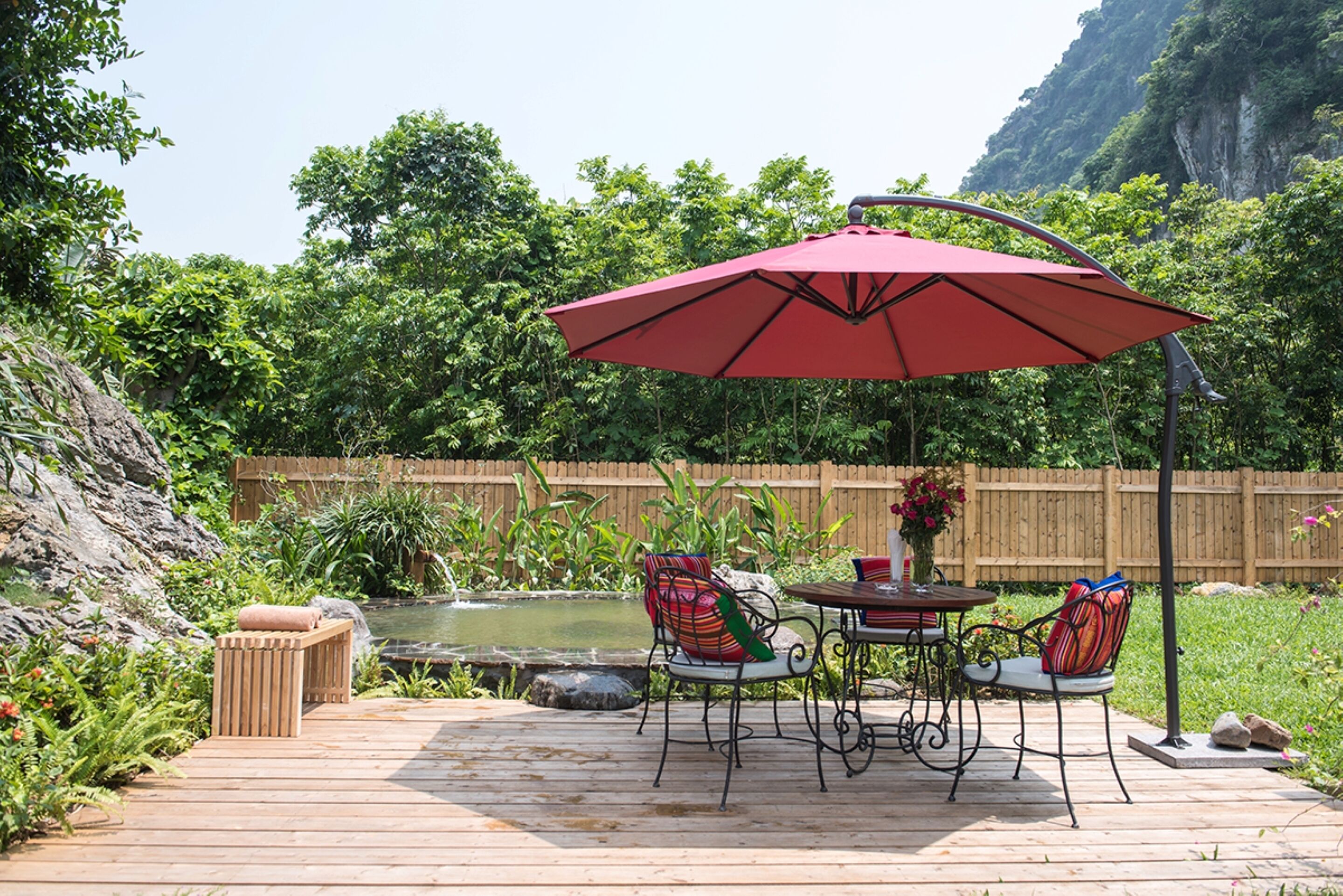 A wooden deck with a table and chairs, a red umbrella, and a small pond.