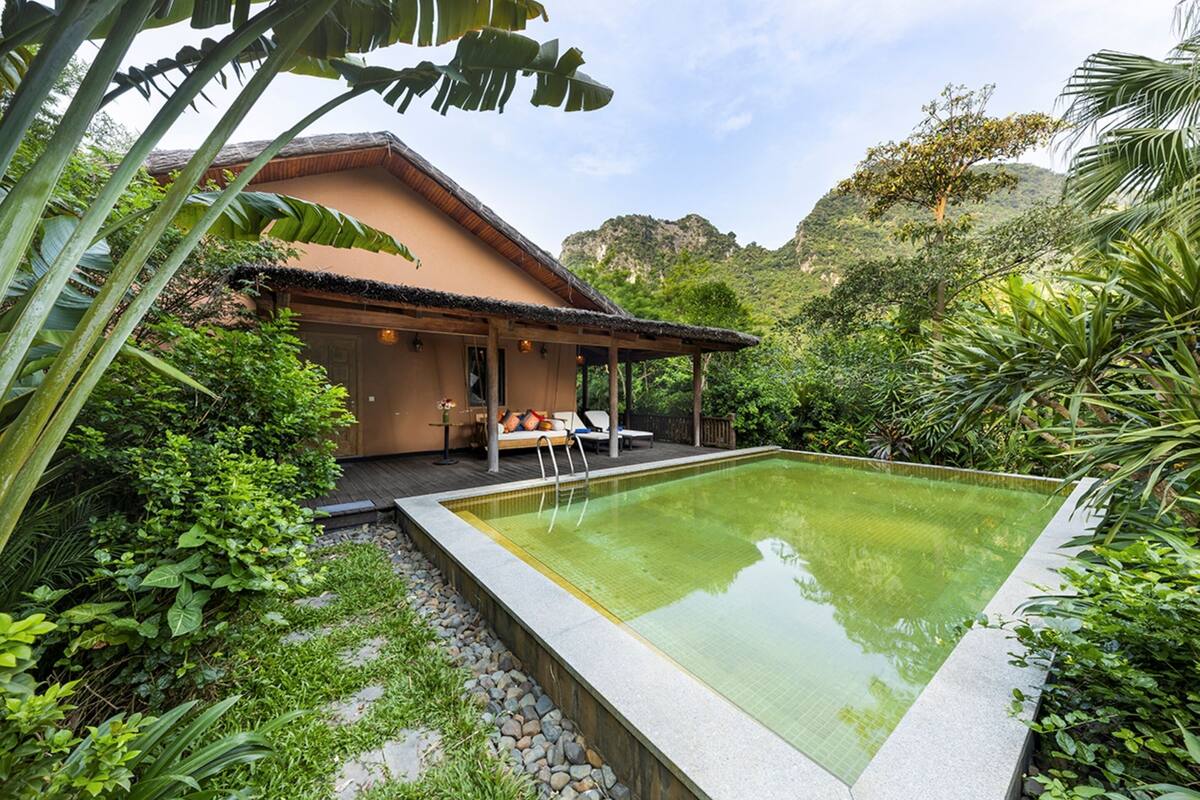 A pool with a wooden deck and lounge chairs, surrounded by lush greenery and a mountain backdrop.