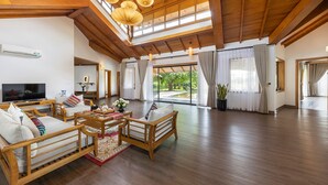 A spacious living room with wooden furniture, a flat-screen TV, and large windows offering a view of greenery.