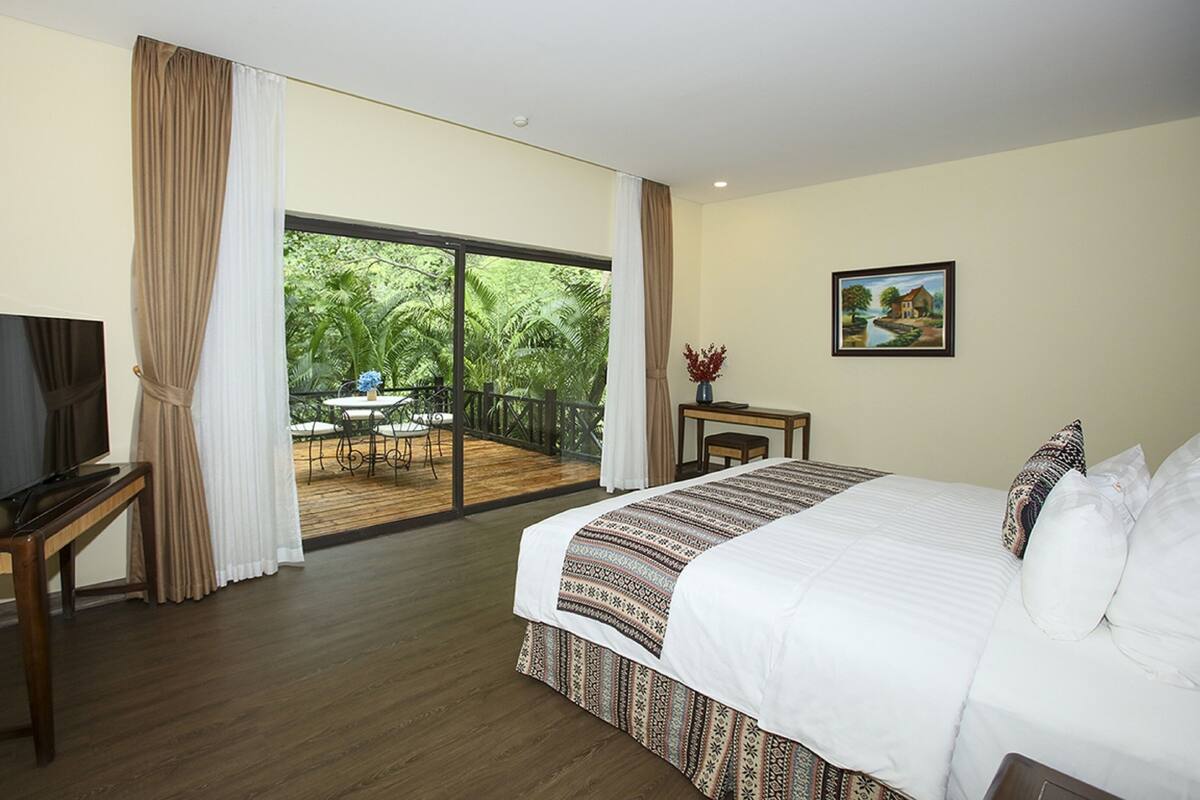 A hotel room with a large bed, a TV, a balcony with a table and chairs, and a painting on the wall.