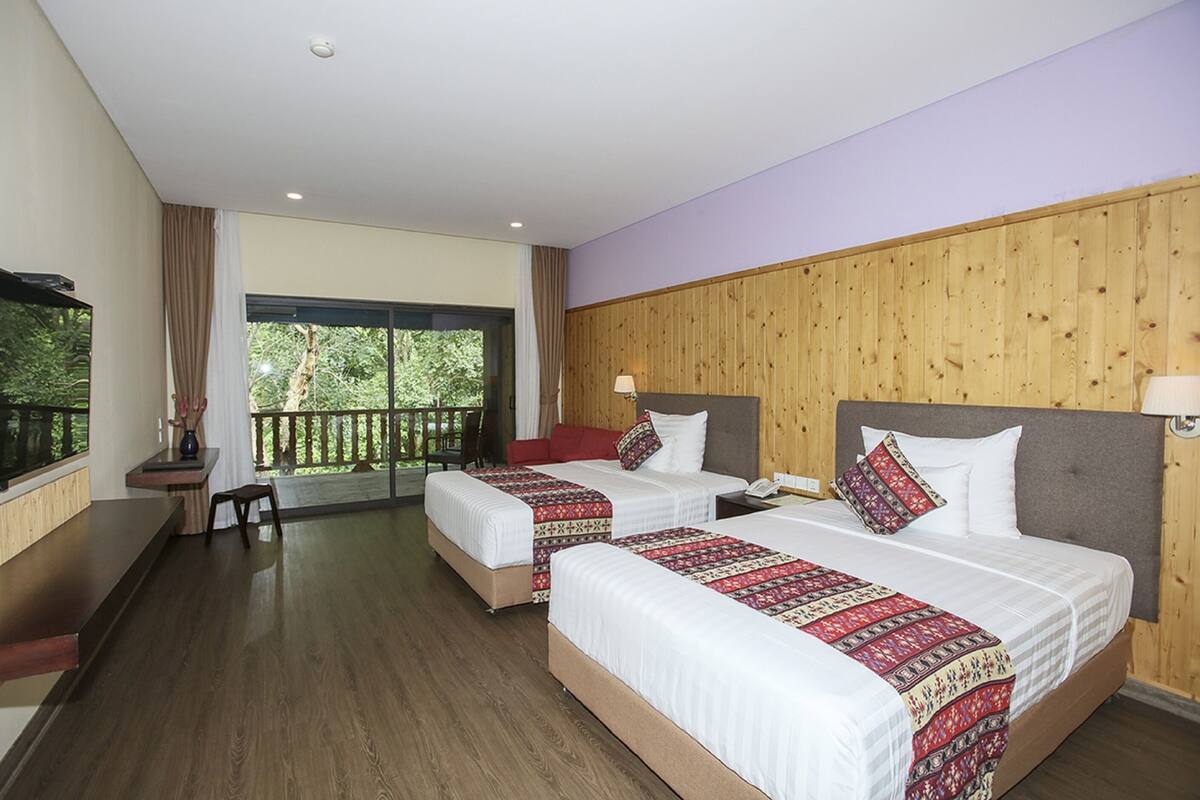 A hotel room with two beds, a desk, a chair, a TV, and a balcony with a view of trees.