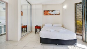 A hotel room with a bed, a small table, a chair, a painting on the wall, and a window with curtains. - Shandelle Apartments (Alexandra Headland)