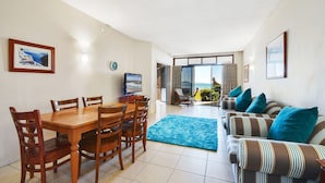 A dining area with a wooden table and chairs, a blue rug, and a striped sofa with teal cushions. - Shandelle Apartments (Alexandra Headland)