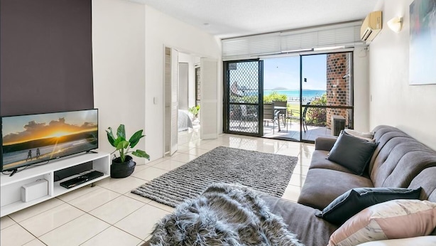A modern living room with a flat-screen TV, a grey sofa, a large rug, and a view of the sea through a sliding glass door. - Shandelle Apartments (Alexandra Headland)
