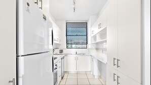 A modern kitchen with white cabinetry, a stainless steel refrigerator, a microwave, and a window with a metal grid. - Shandelle Apartments (Alexandra Headland)