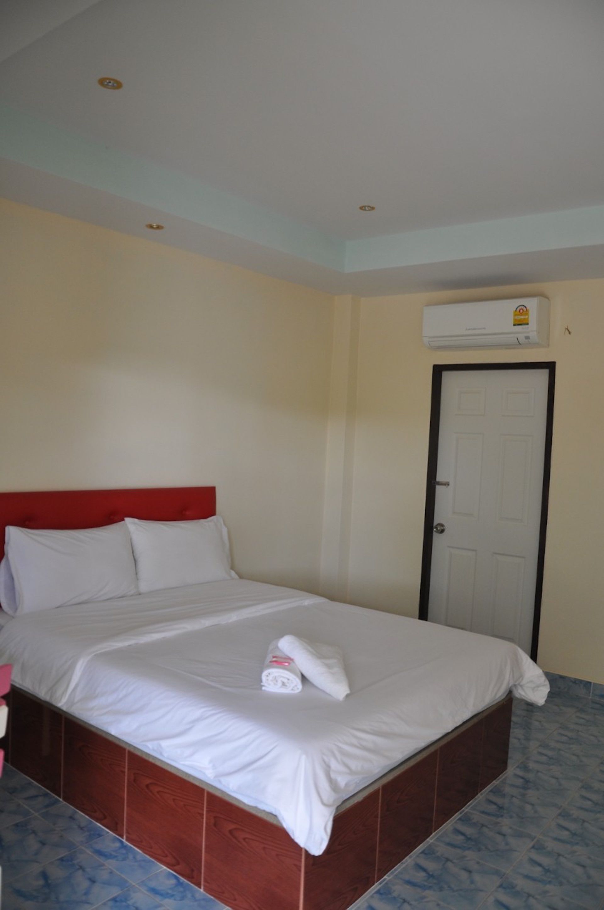 A hotel room with a single bed, a red headboard, white bedding, a white door, and an air conditioning unit on the wall.