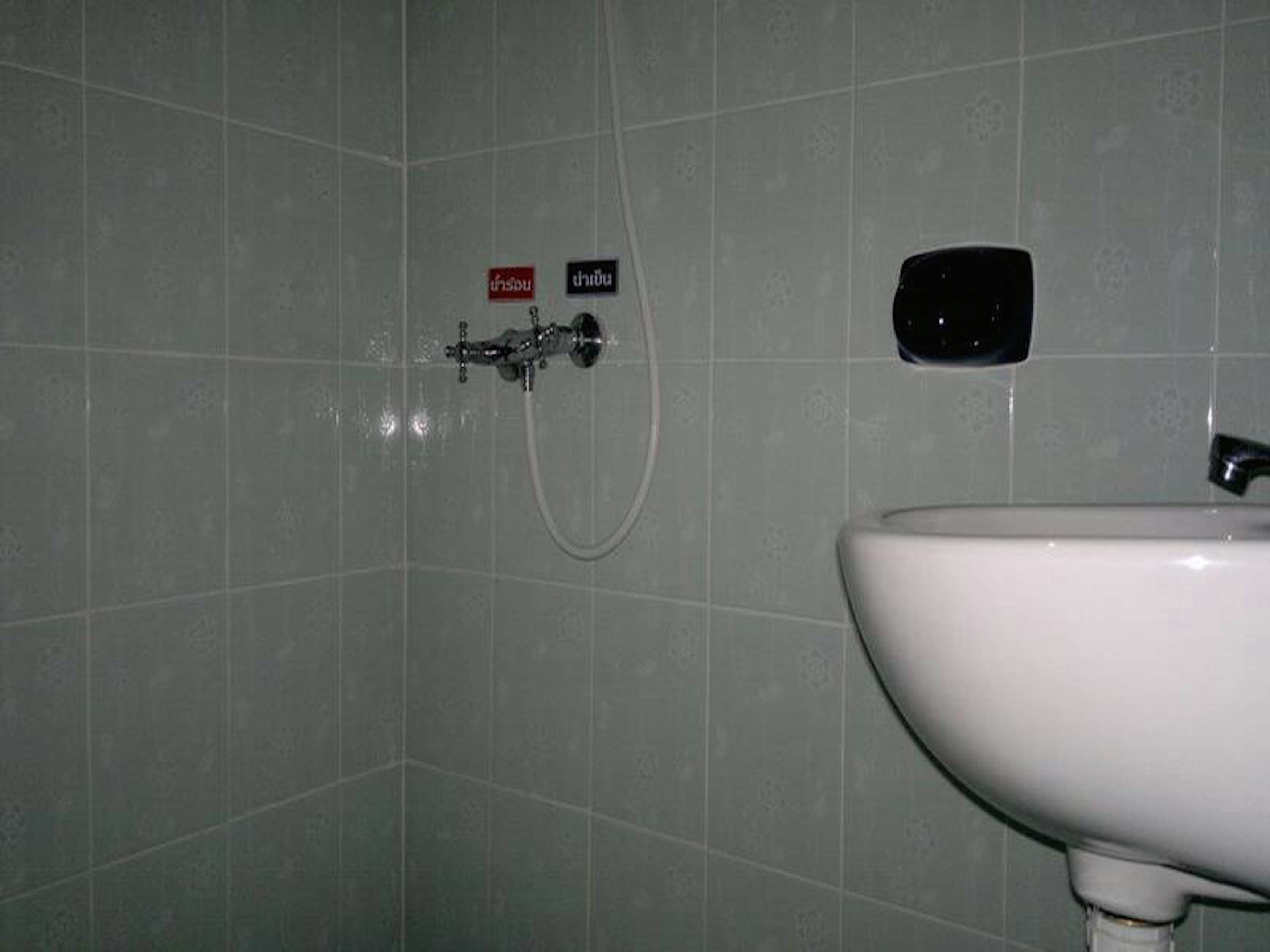 Ein Badezimmer mit einem weißen Waschbecken, einer Dusche mit Bedienfeld und einem an der Wand befestigten Schild mit thailändischem Text.