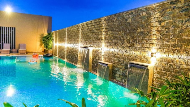 A swimming pool with a stone wall and waterfalls, surrounded by lounge chairs and potted plants. - Vivera Grande (Thindukkal)