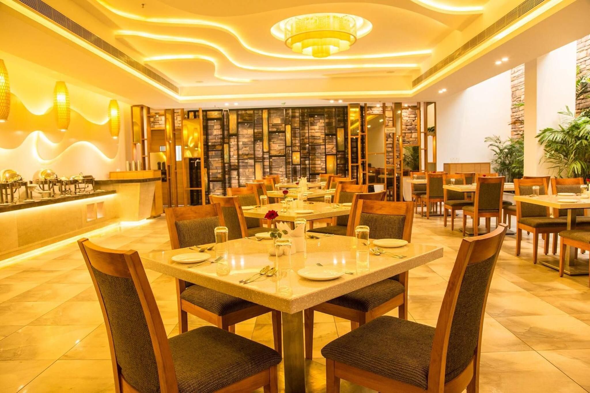 A restaurant with multiple dining tables and chairs, a central chandelier, and a stone wall backdrop.