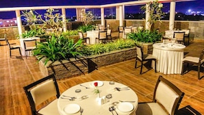 An outdoor dining area with round tables and chairs, surrounded by potted plants and a city view at night. - Vivera Grande (Thindukkal)