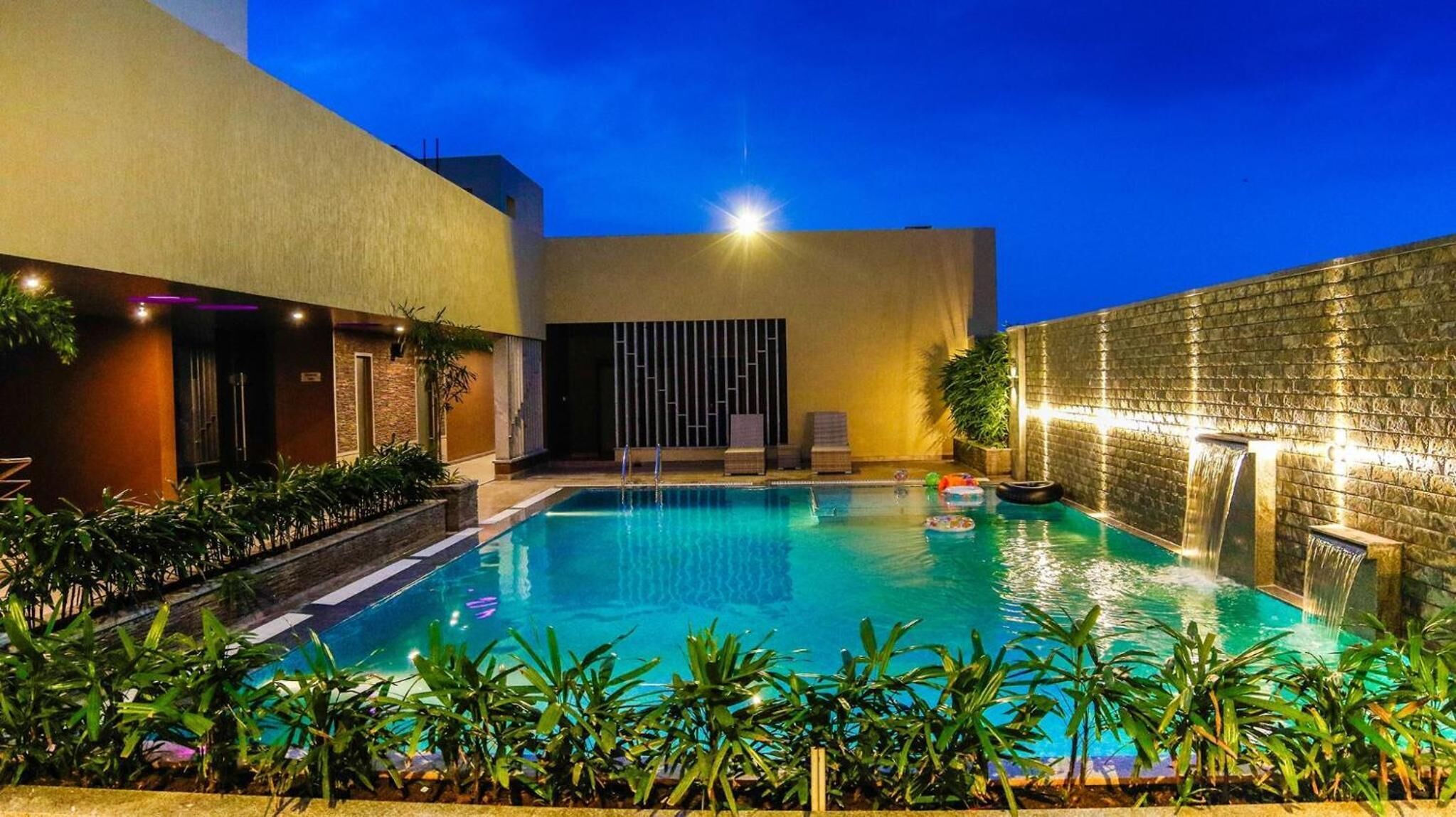 A swimming pool with a waterfall feature, surrounded by plants and a modern building facade.