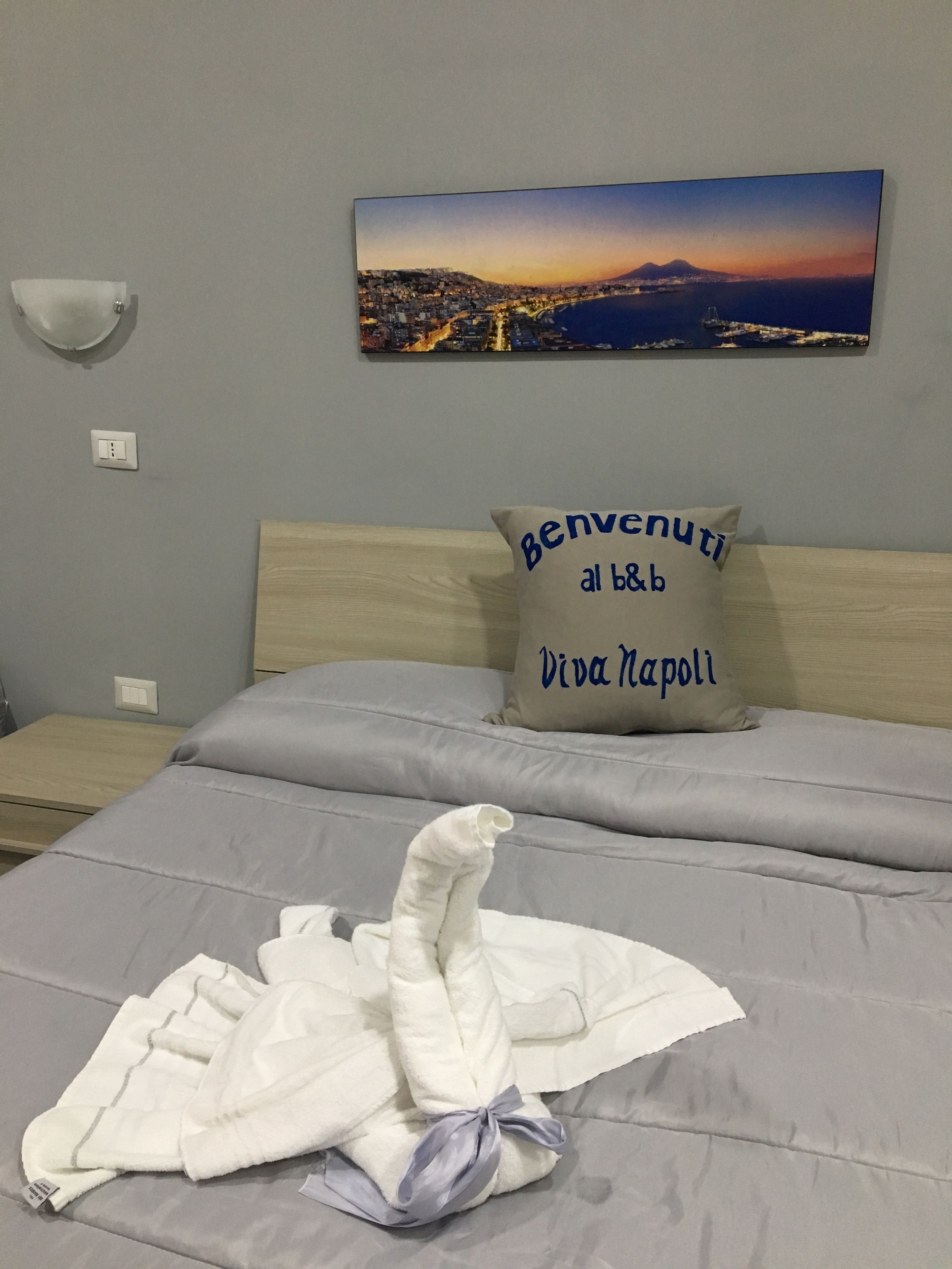 Un lit bien fait, agrémenté d’une serviette et d’un peignoir, un oreiller avec l’inscription « Bienvenue au B&B Viva Napoli » et un tableau représentant un paysage urbain accroché au mur.