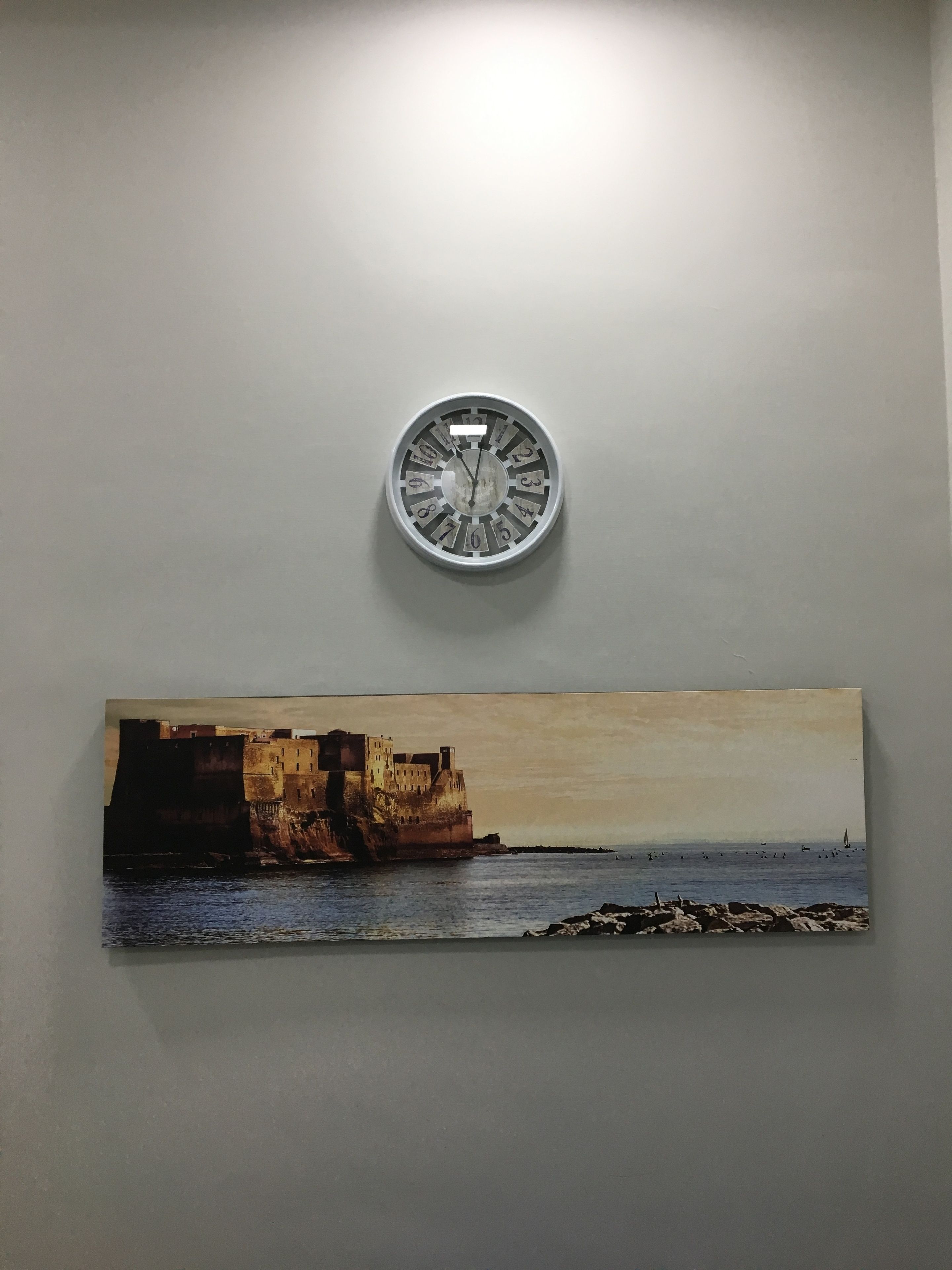A wall-mounted clock with a circular face and numbers, and a painting of a coastal scene with buildings and water.