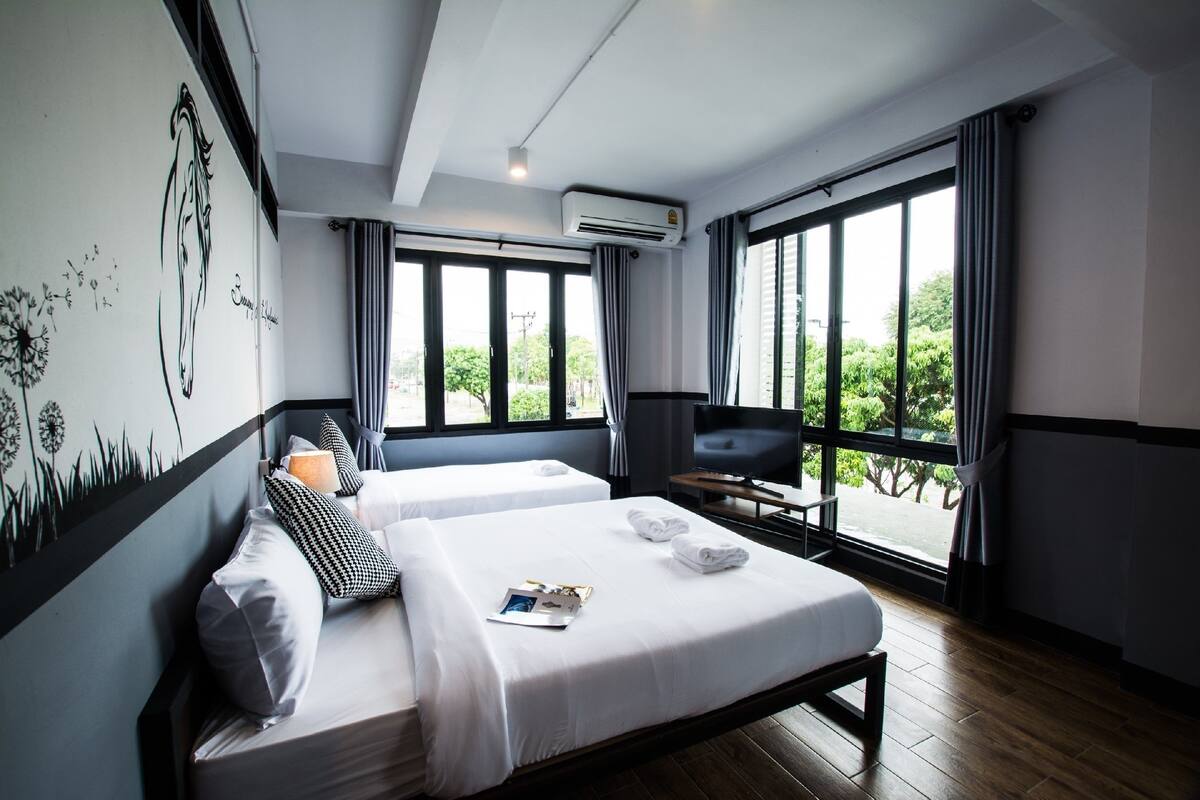 A modern hotel room with two beds, a TV, and large windows offering a view of greenery.