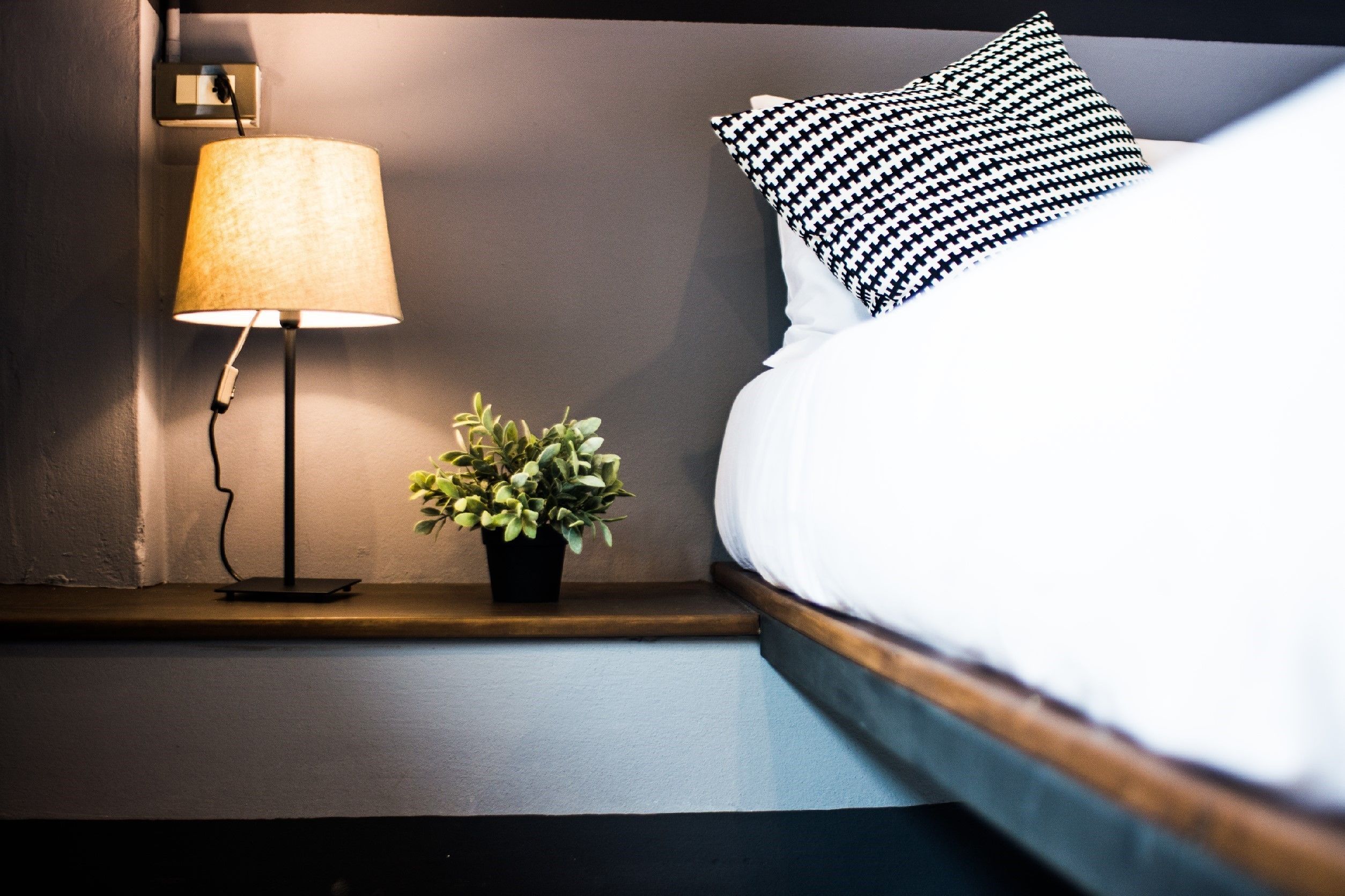 A neatly made bed with a black and white patterned pillow, a bedside lamp, and a small potted plant on a bedside table.