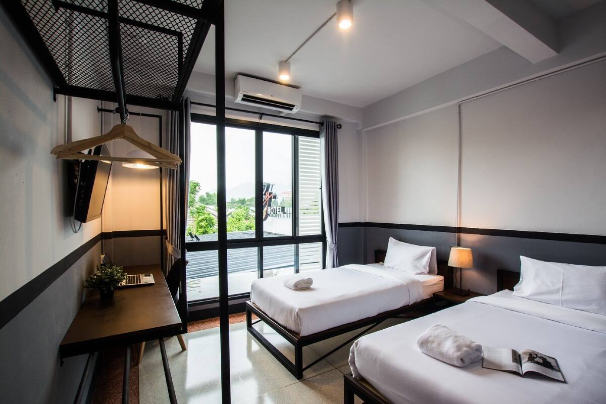A modern hotel room with two beds, a desk, and a large window offering a view of the outdoors.