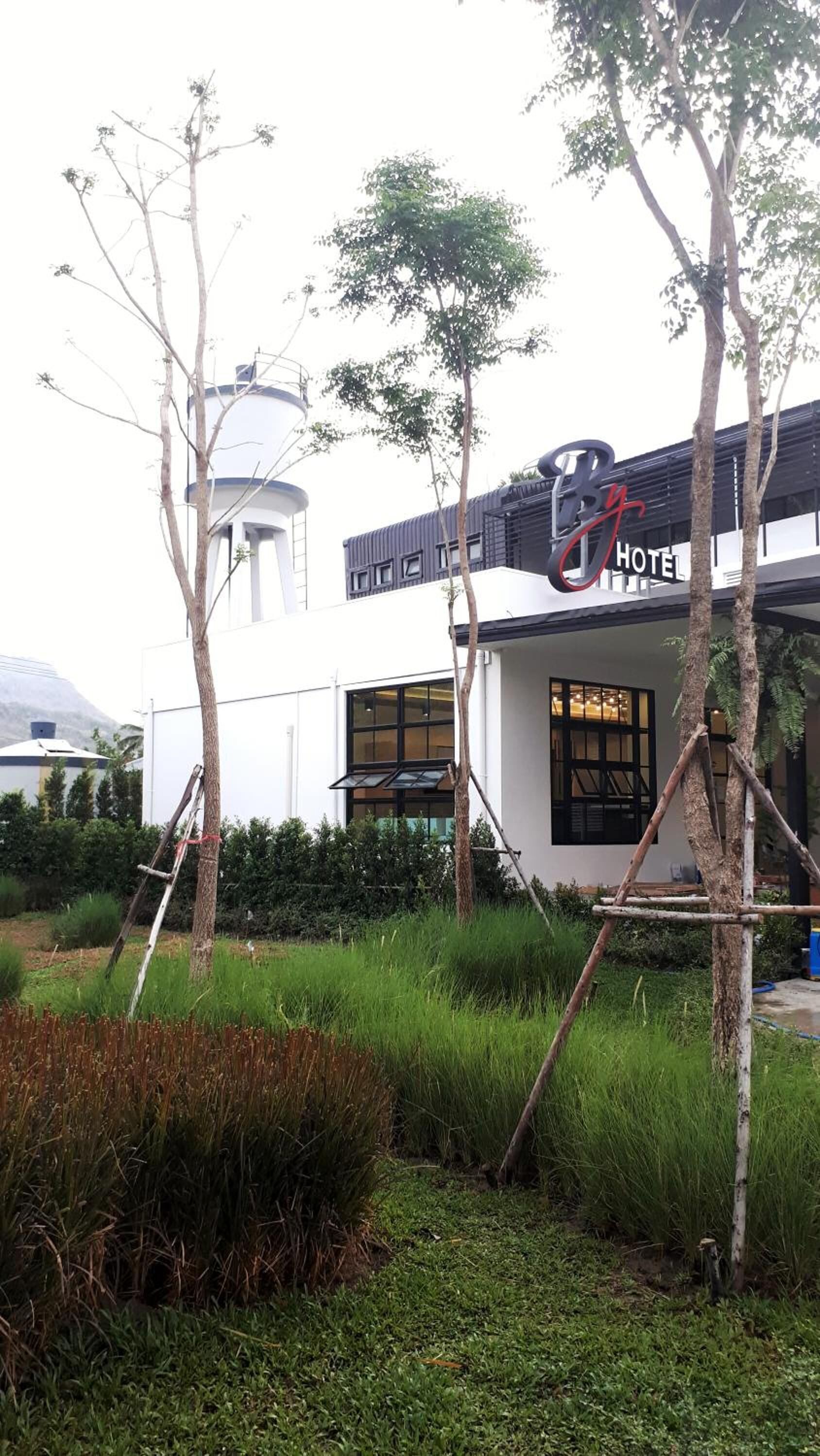 A hotel with a modern design, featuring a prominent 'By Hotel' sign, a water tower in the background, and a well-maintained garden with trees and grass.