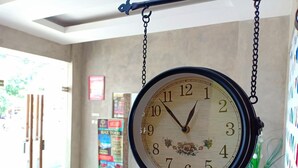 A large analog clock hanging from the ceiling in a room with a tiled floor, a wooden bench, and a poster on the wall. - Hazotel Semarang (Semarang)