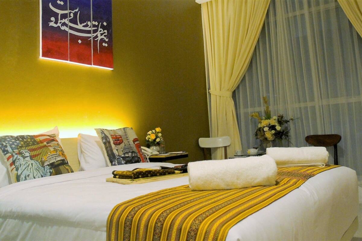A hotel room with a bed, pillows, a nightstand, a lamp, and a window with curtains.