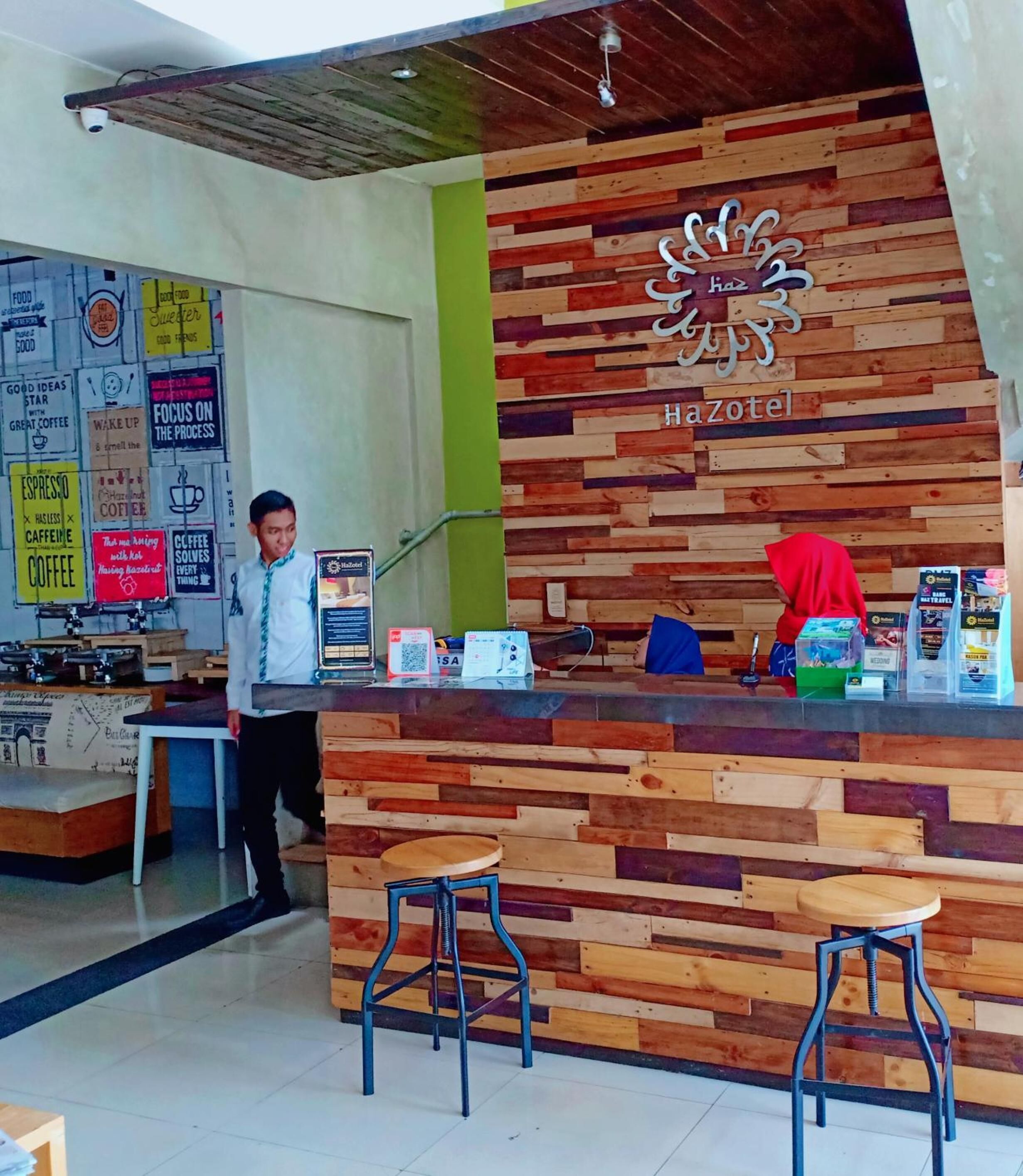 A wooden reception desk with a sign reading "HaZotel" and a man standing behind it. There are two stools in front of the desk.