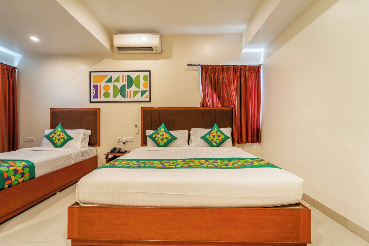 A hotel room with two beds, a large window with red curtains, an air conditioning unit, and a colorful abstract painting on the wall.