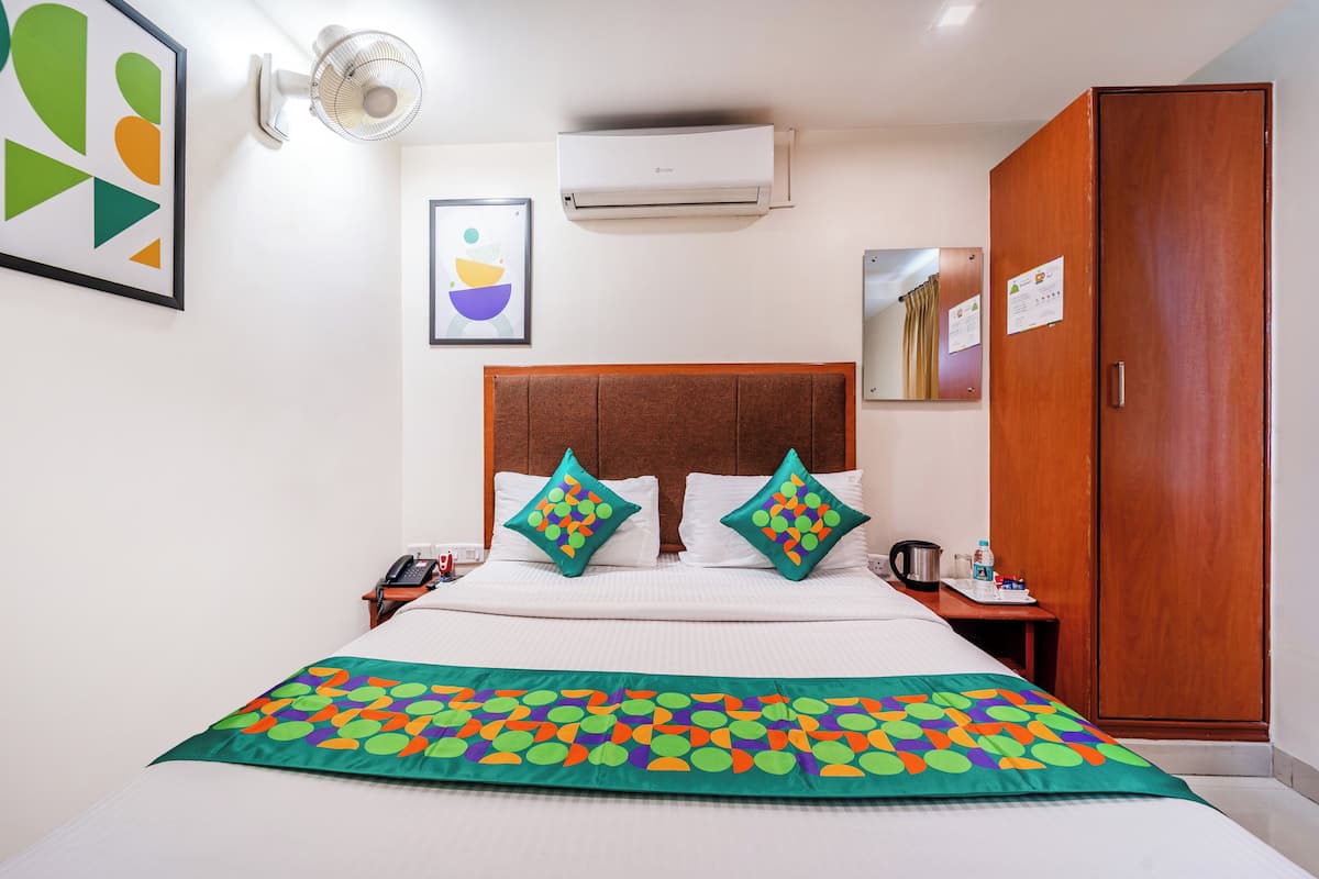A hotel room with a bed, a bedside table with a phone, a coffee maker, a cabinet, and an air conditioning unit.