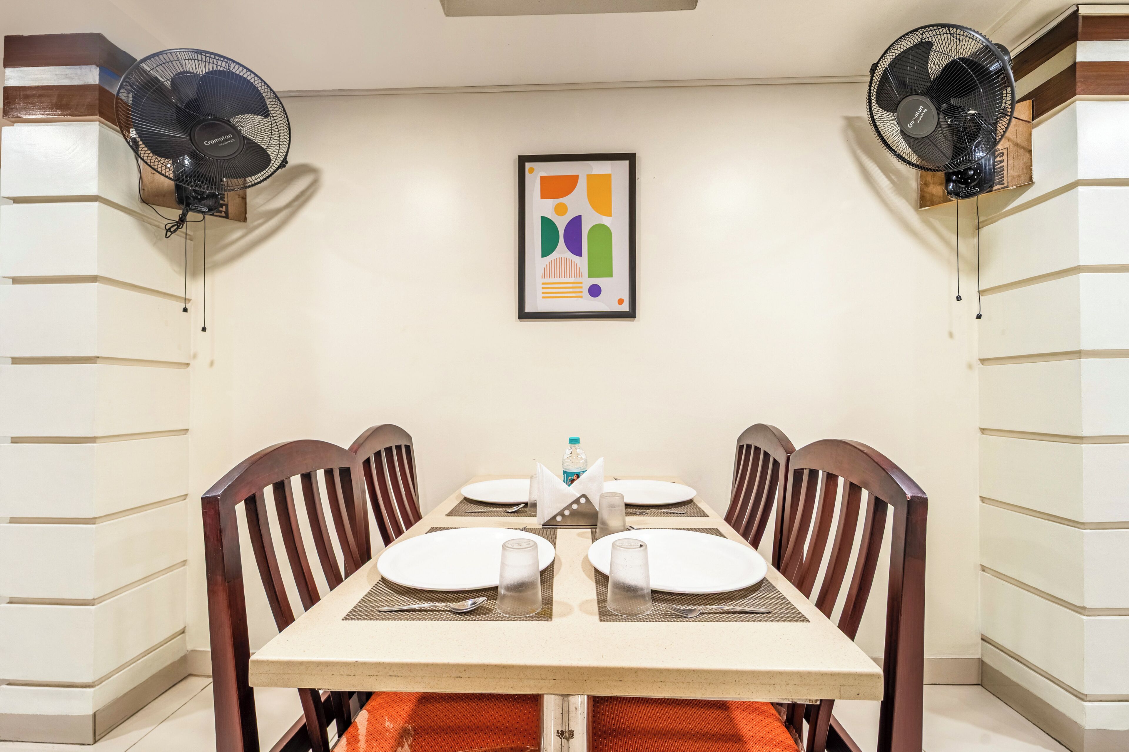 Un comedor con una mesa dispuesta para cuatro, dos ventiladores de pared y una obra de arte abstracta enmarcada en la pared.