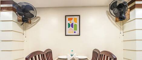 Un comedor con una mesa dispuesta para cuatro, dos ventiladores de pared y una obra de arte abstracta enmarcada en la pared.