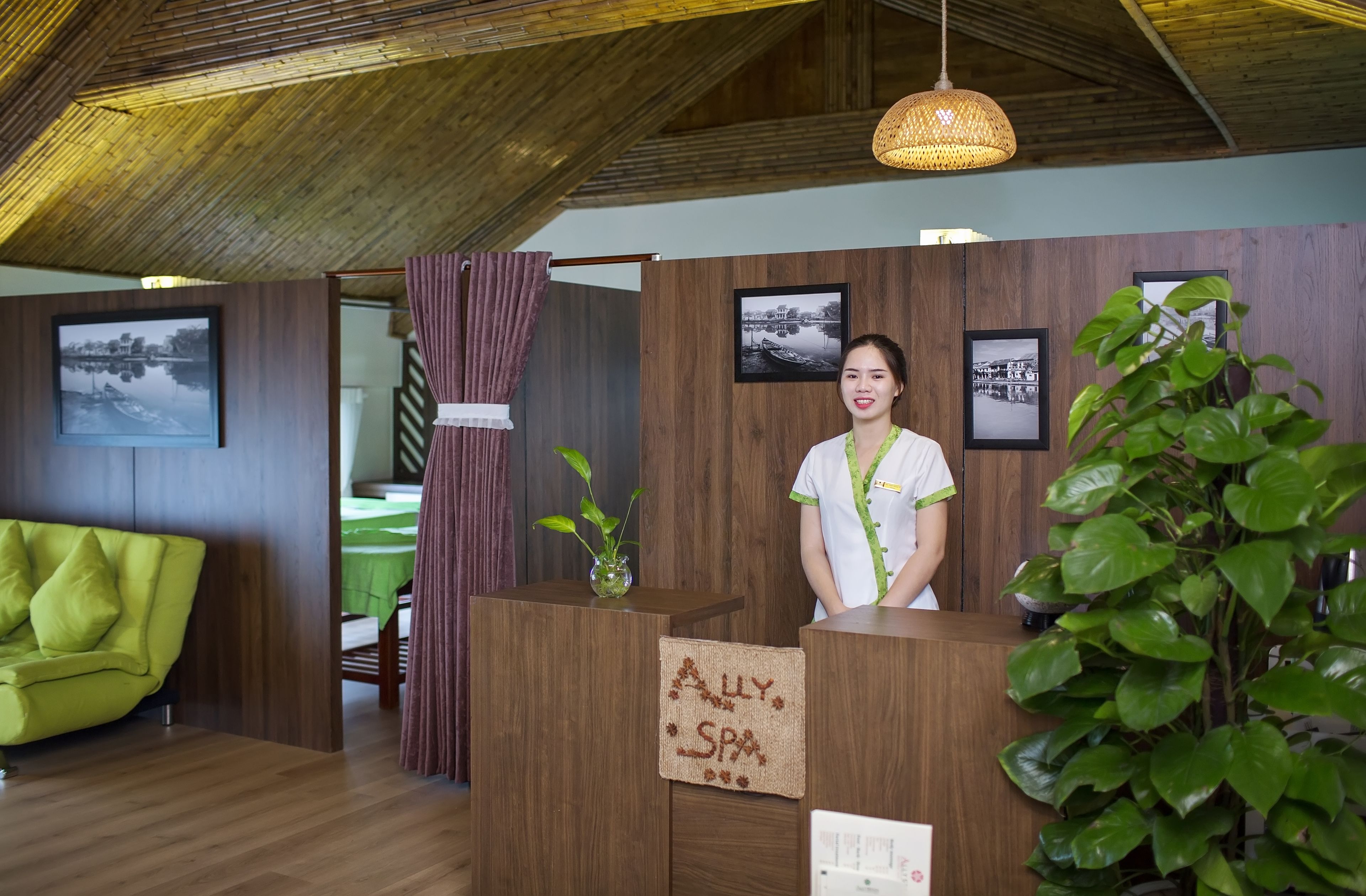 Un comptoir d’accueil avec une enseigne indiquant « ALLY SPA », situé dans une pièce aux murs en bois, avec un canapé vert et un luminaire suspendu.