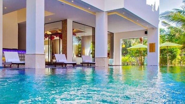 A modern hotel pool area with clear blue water, sun loungers, and large windows offering views of the surrounding greenery. - Ally Beach Boutique Hotel Hoian (Da Nang)