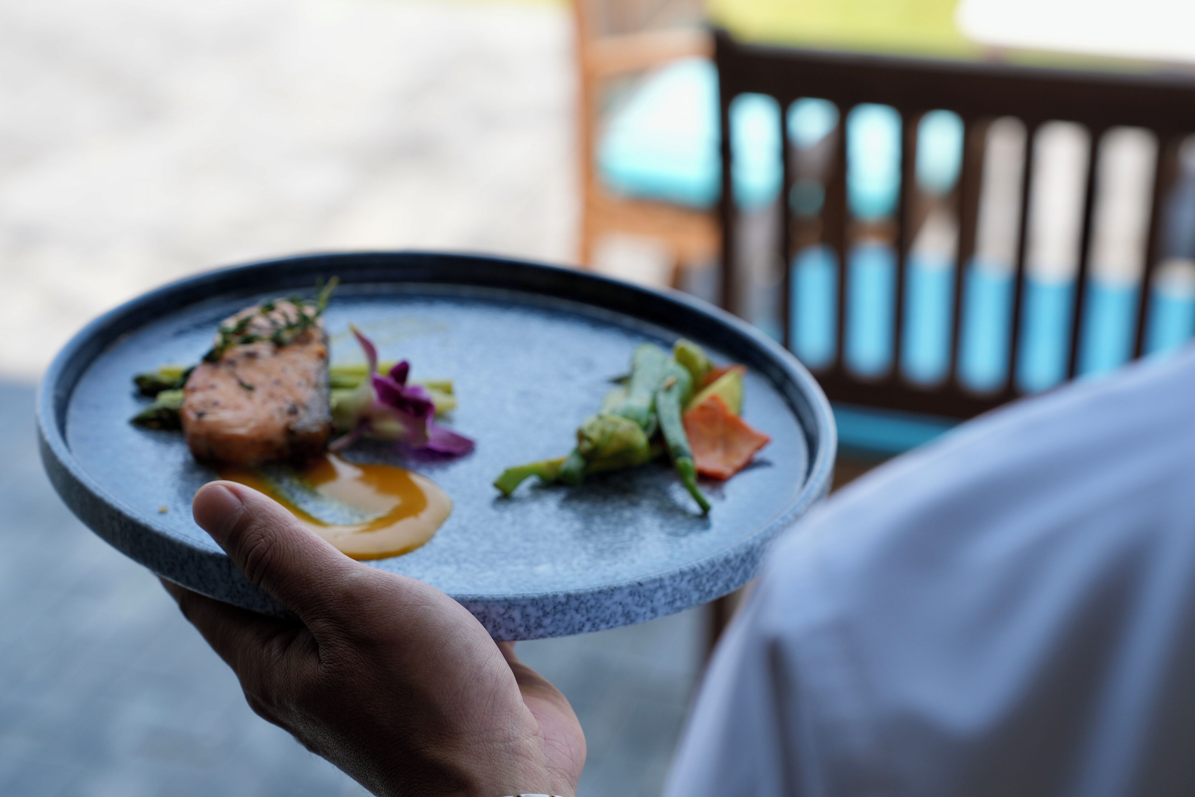 A person holding a plate with food, a chair, and a table in the background.