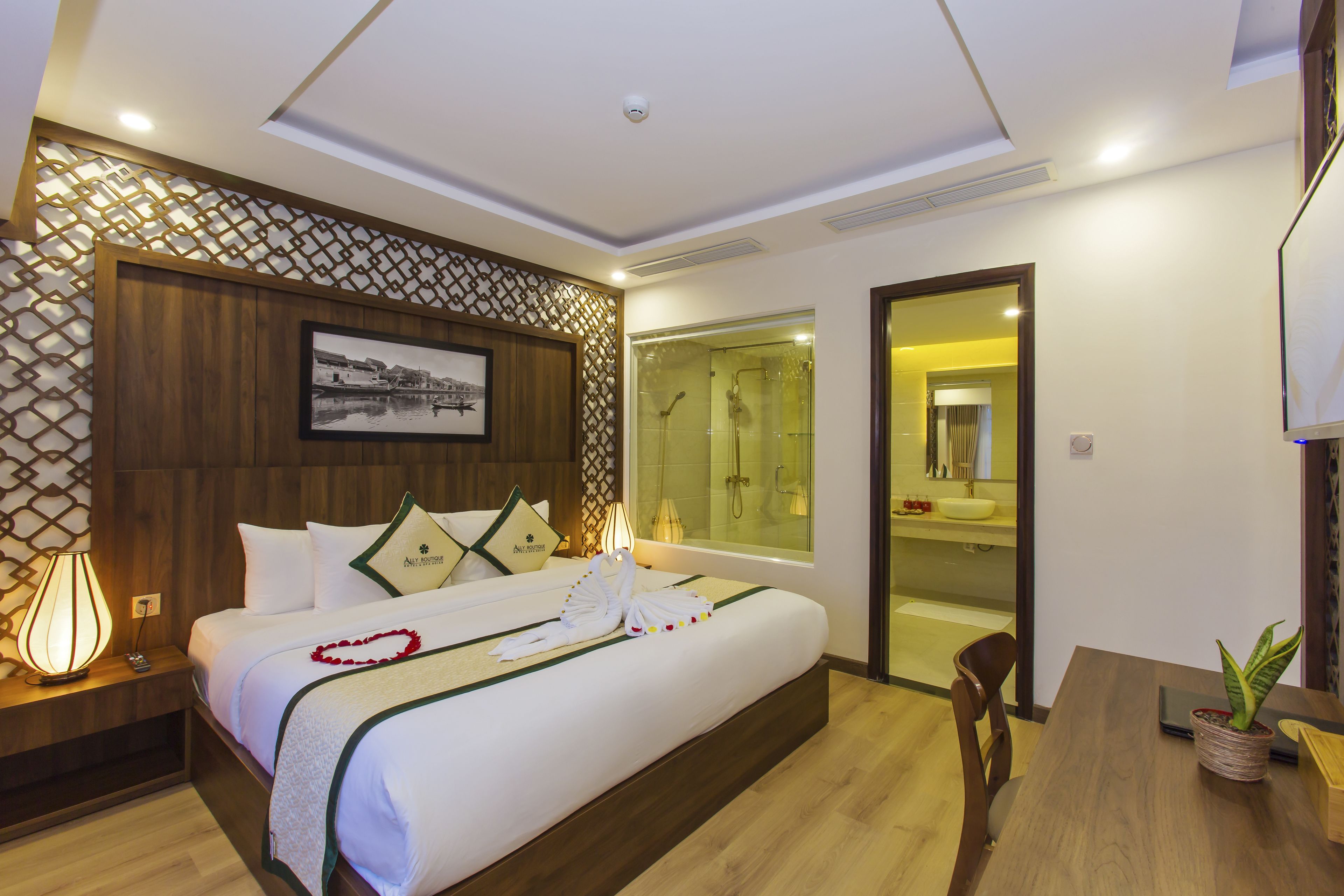 Habitación de hotel con una cama grande, una mesita de noche con lámpara, cabecero de madera, un cuadro enmarcado en la pared y una puerta corredera que da a un baño.