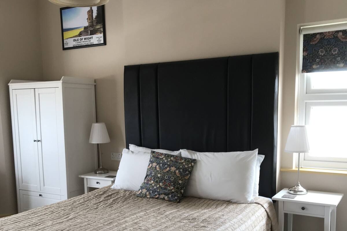 A neatly arranged bedroom with a large bed, a dark headboard, a white wardrobe, and a framed picture on the wall.