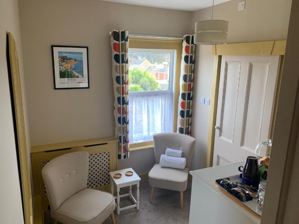 A small, cozy room with a window, a chair, a small table, and a kitchenette area.