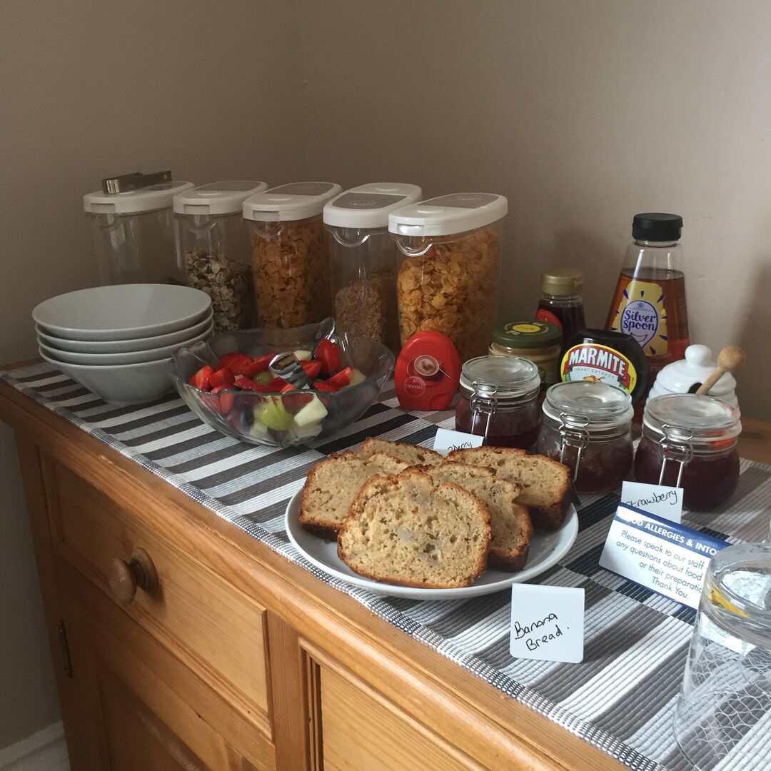 A breakfast buffet with sliced banana bread, jars of marmite, and a bowl of fruit.