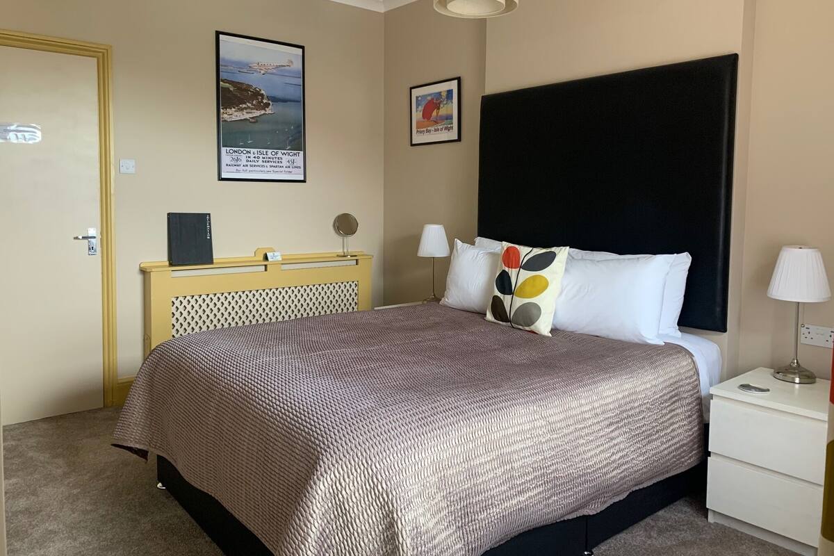 A hotel room with a bed, a nightstand, a lamp, a framed picture of a boat, and a sign about the London & Isle of Wight route.