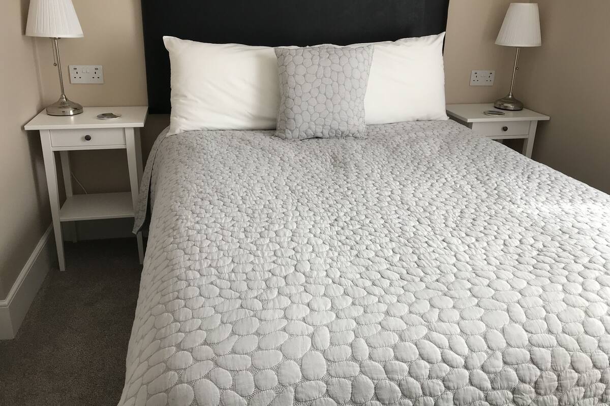 A neatly made bed with a textured white duvet, two bedside tables with lamps, and a plain wall.