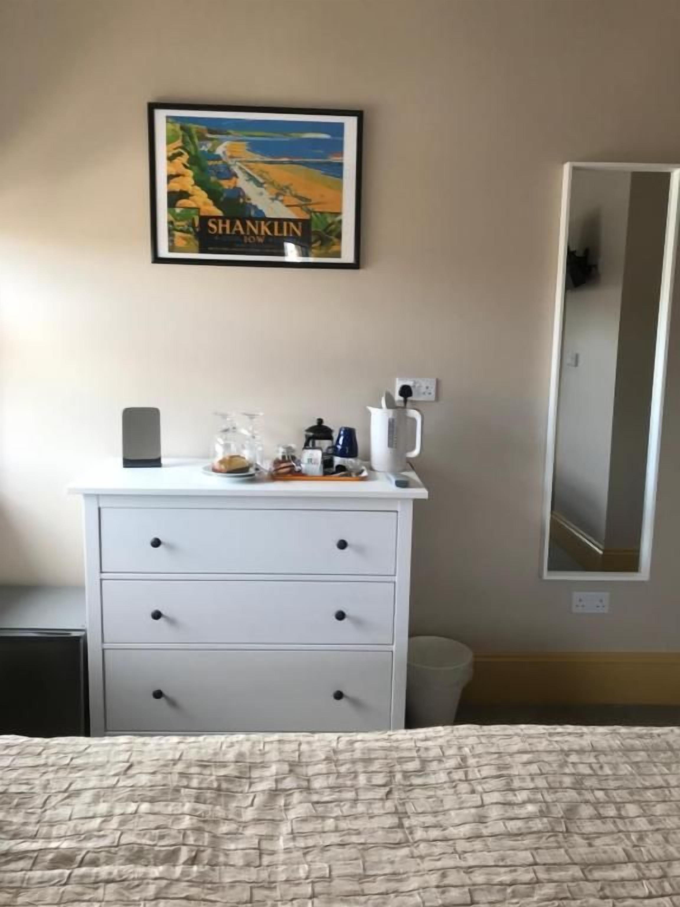 A hotel room with a bed, a dresser with drawers, a framed picture on the wall, a mirror, and a nightstand with a lamp and a coffee maker.