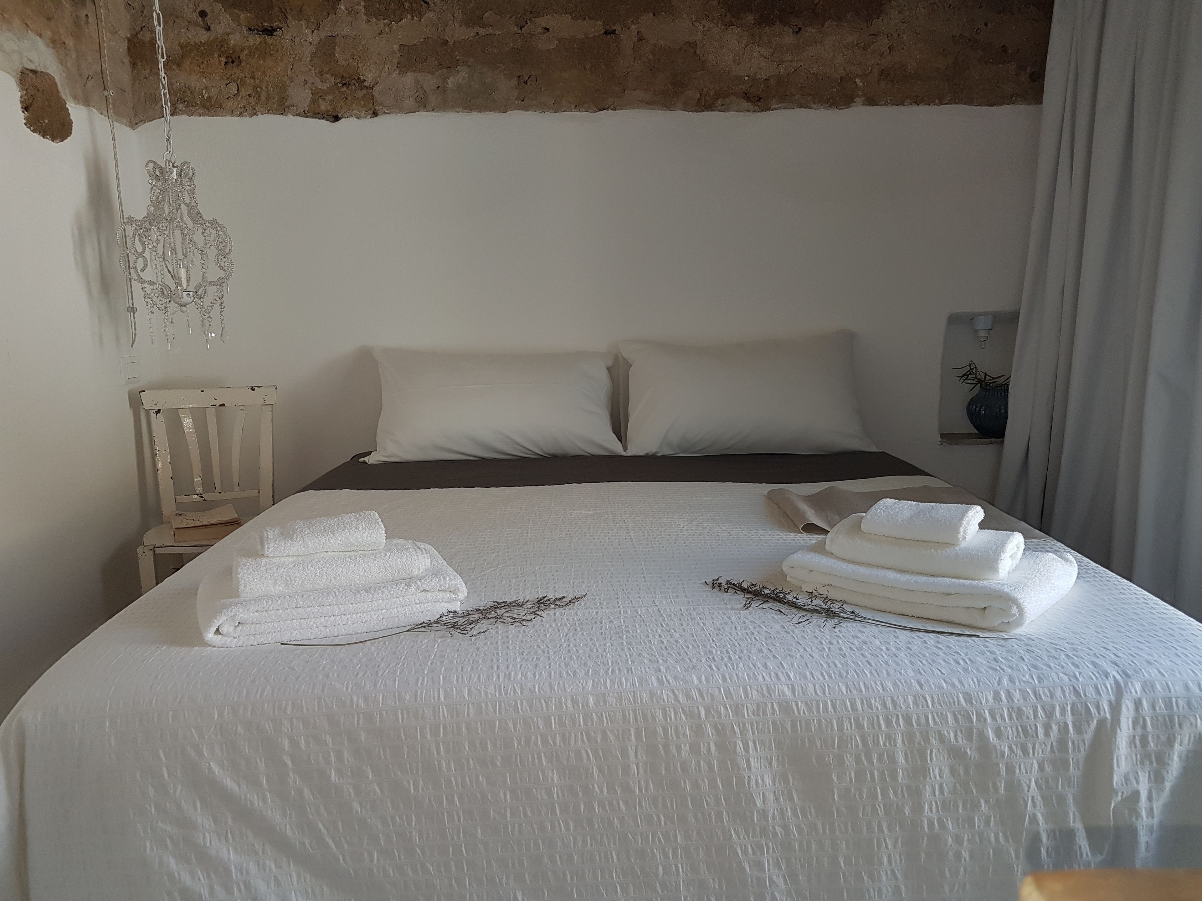 A neatly made bed with white linens and towels, a rustic brick wall, and a simple bedside table with a lamp.